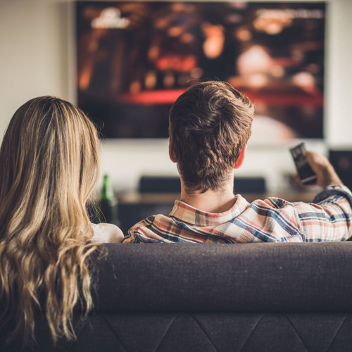 The back of a couple's heads while they sit on a couch watching a TV on the wall in front of them